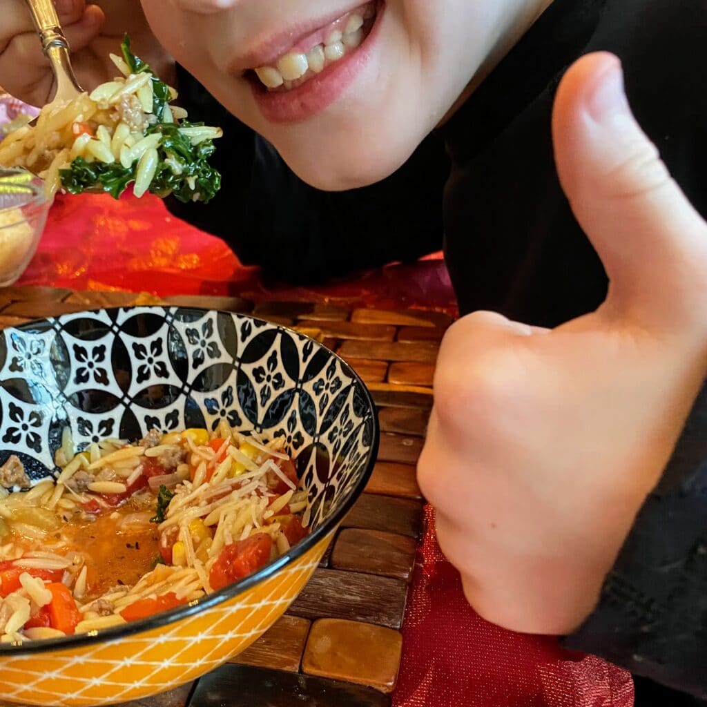 Child smiling and eating sausage orzo soup with kale