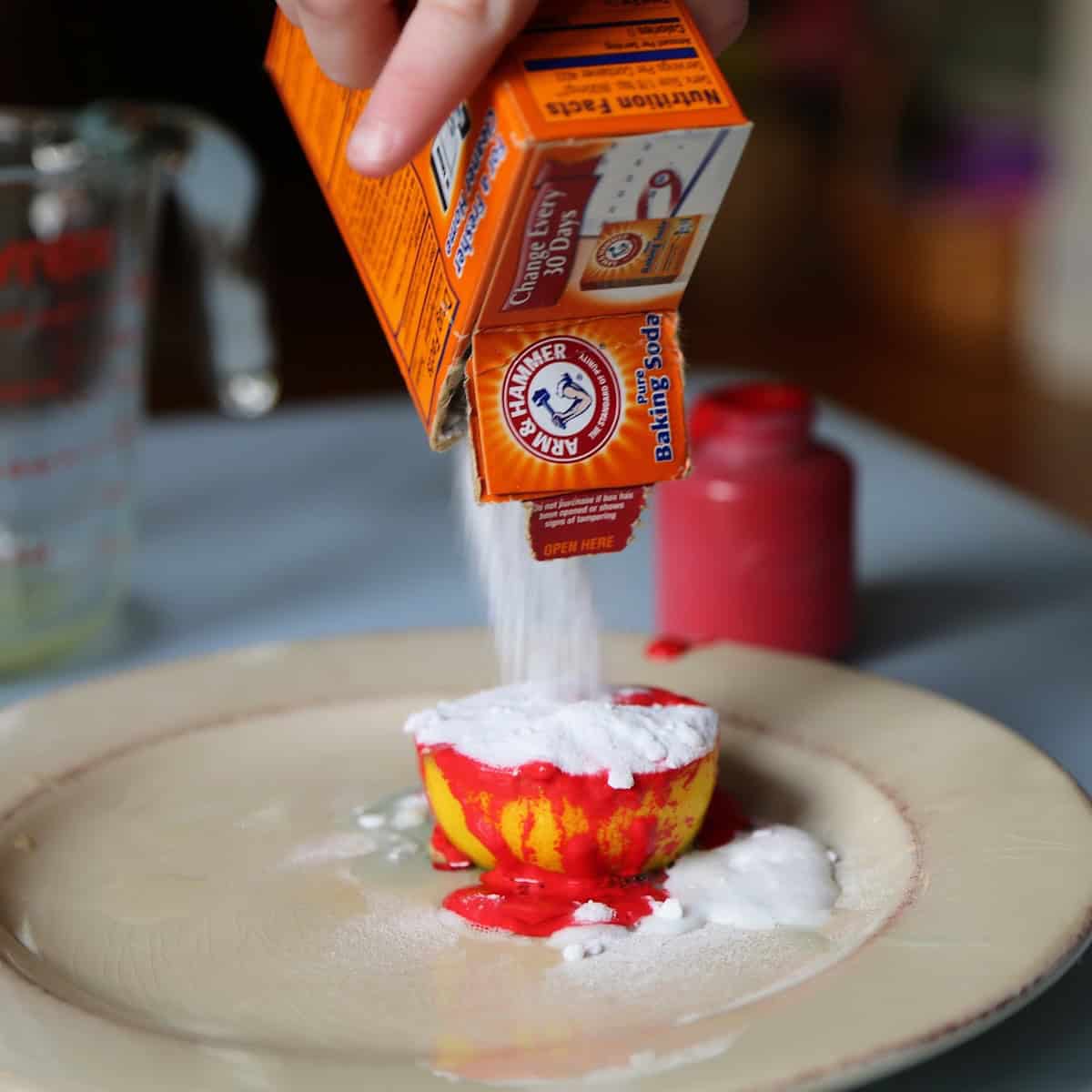 Hand pouring baking soda on top of a halved lemon with red paint. 