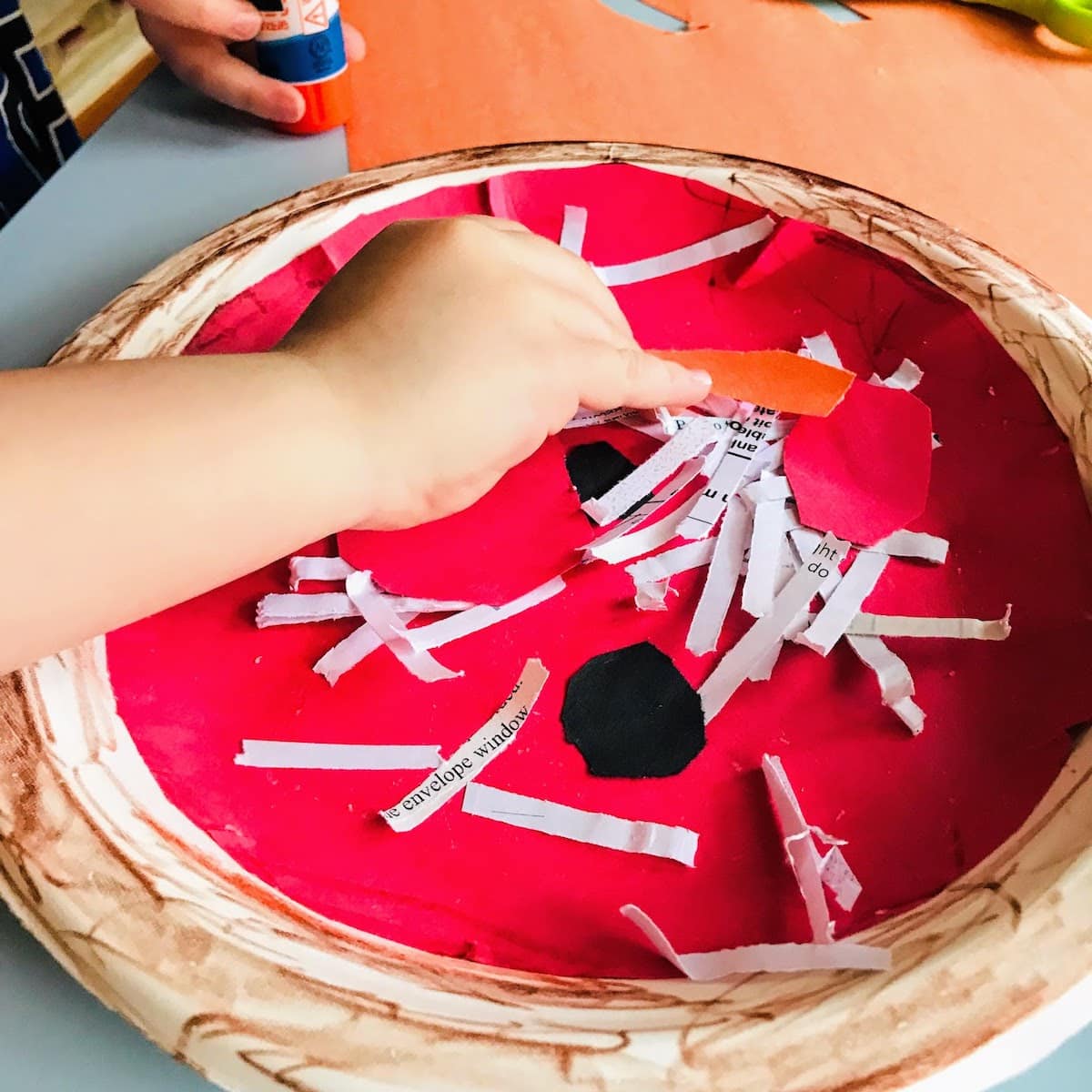 child gluing paper toppings to paper plate pizza