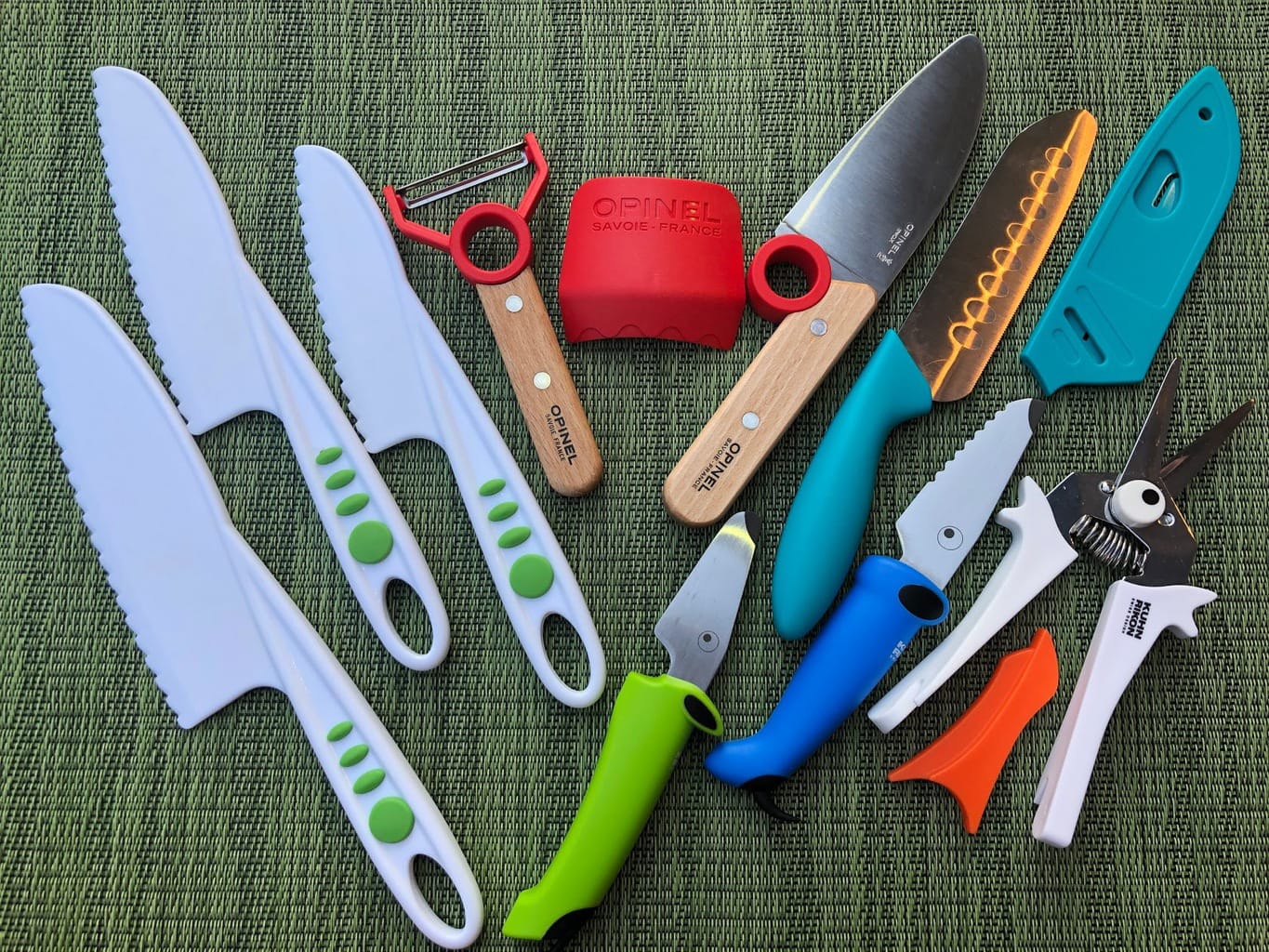 different brands of kids kitchen knives, vegetable peelers, and snippers laid out on a green placement