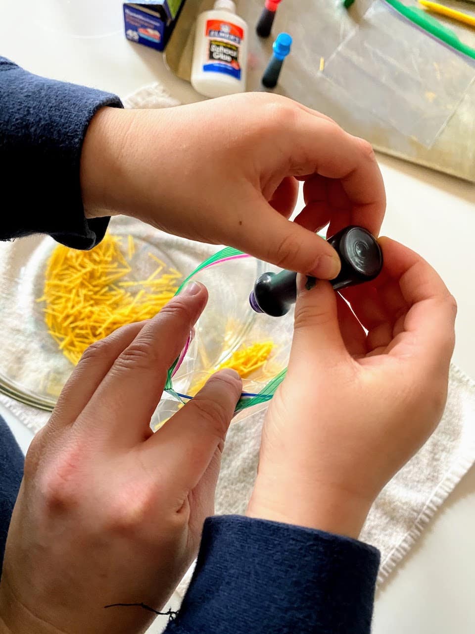 child and parent putting drops of food dye into ziploc bag of broken spaghetti noodles