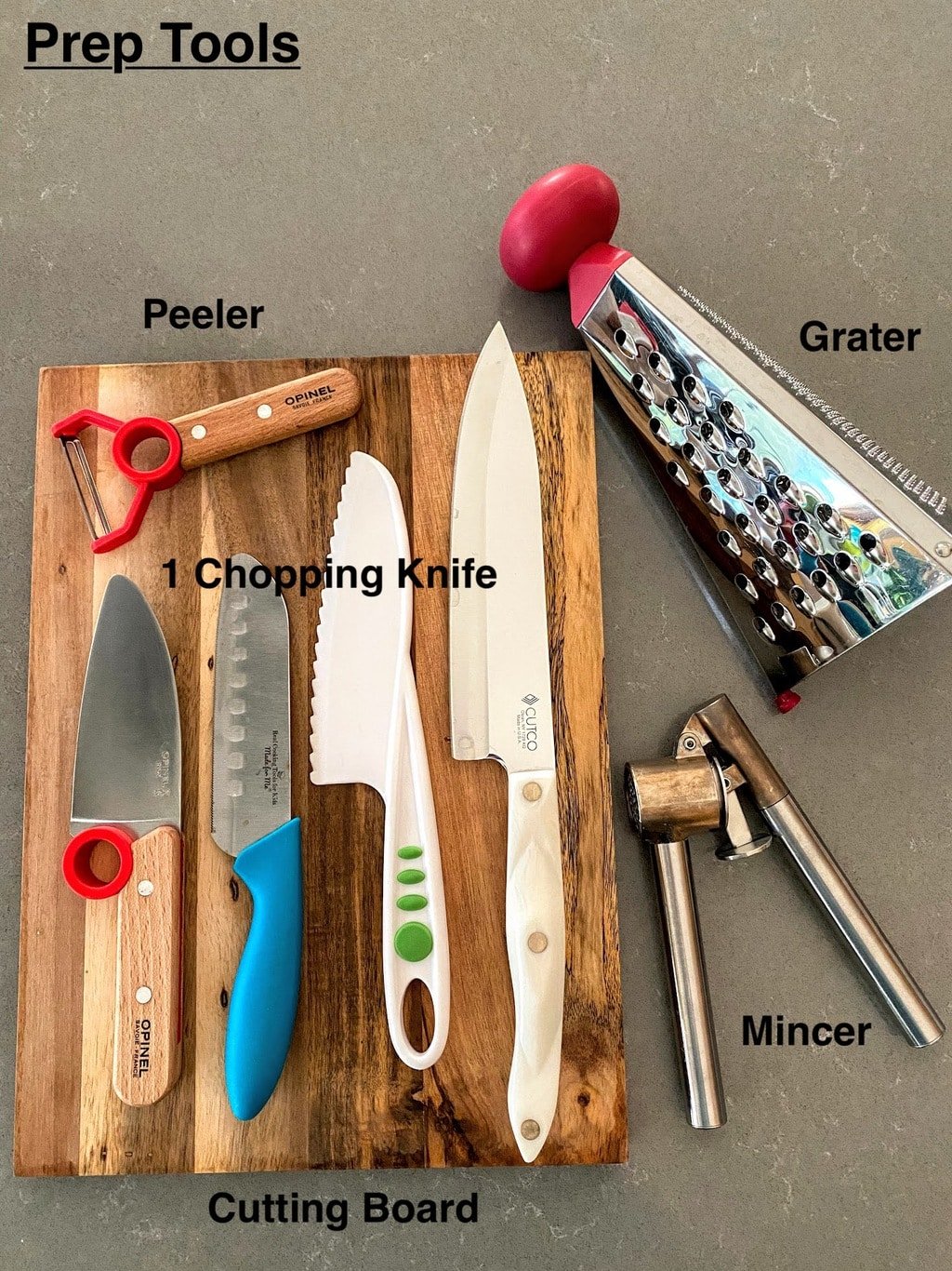 peeler, chopping knives, cutting board, grater and mincer on grey counter