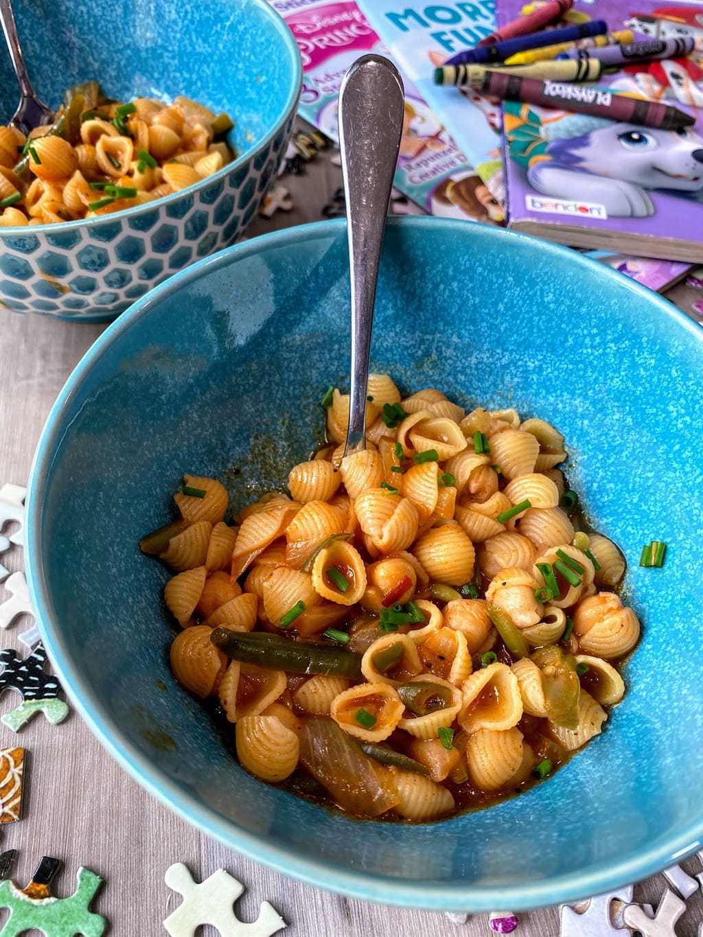 pasta with chickpeas and chives in blue bowl on table with puzzle pieces, crayons and coloring books