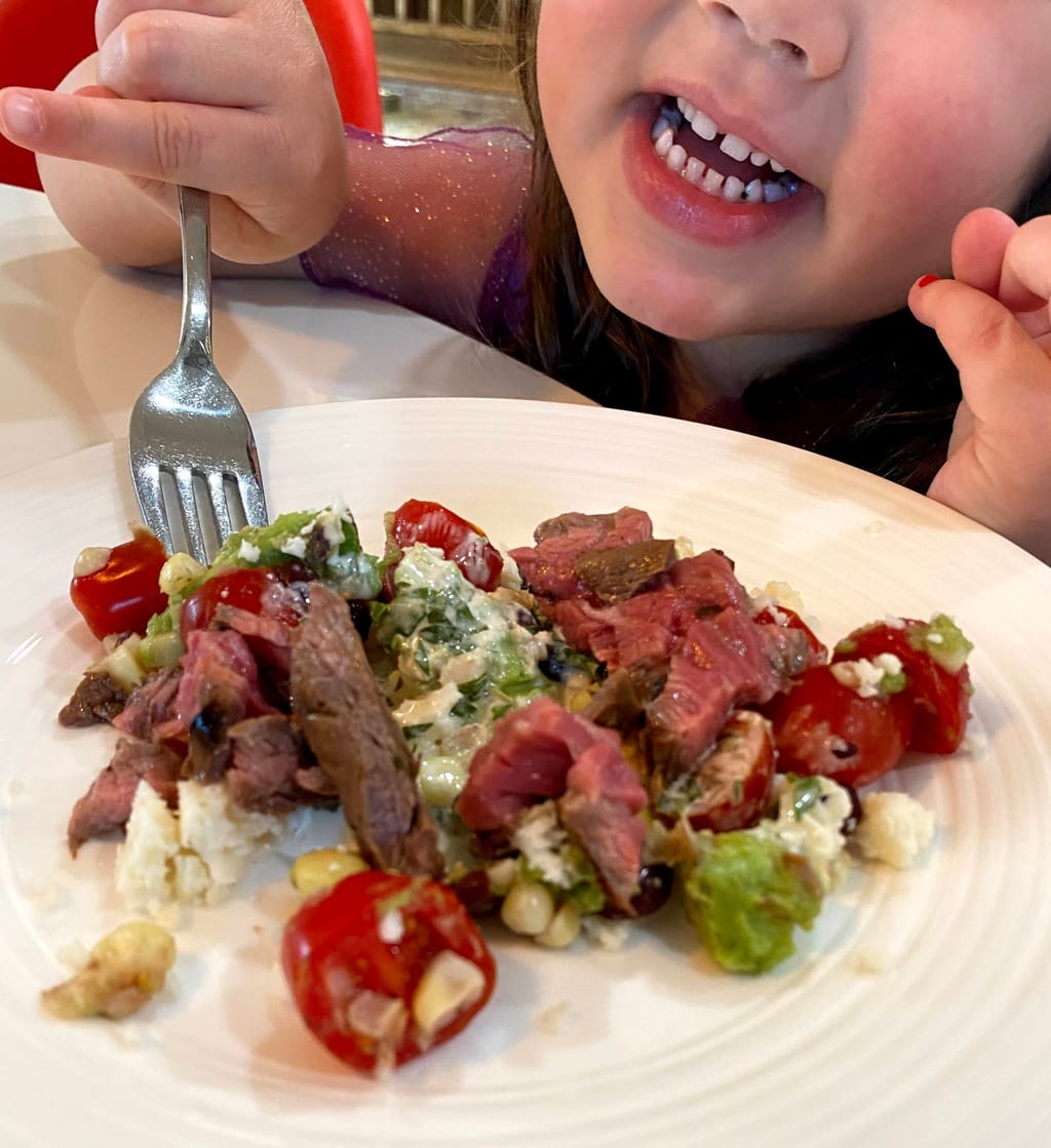 smiling child holding fork over plate of carne asada and vegetables