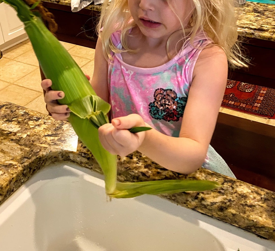 blonde child standing over kitchen sink shucking an ear of corn