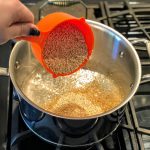 person pouring quinoa into boiling pot of water from orange cup