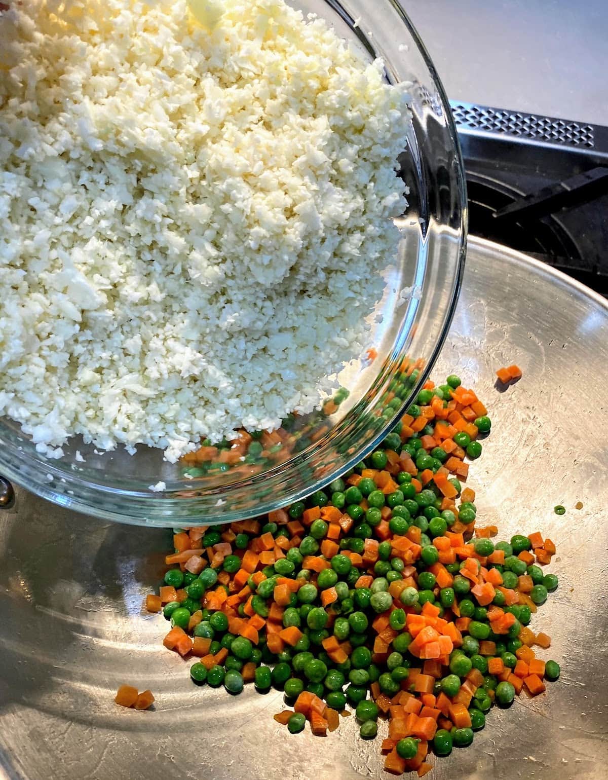 adding riced cauliflower to wok filled with frozen peas and carrots