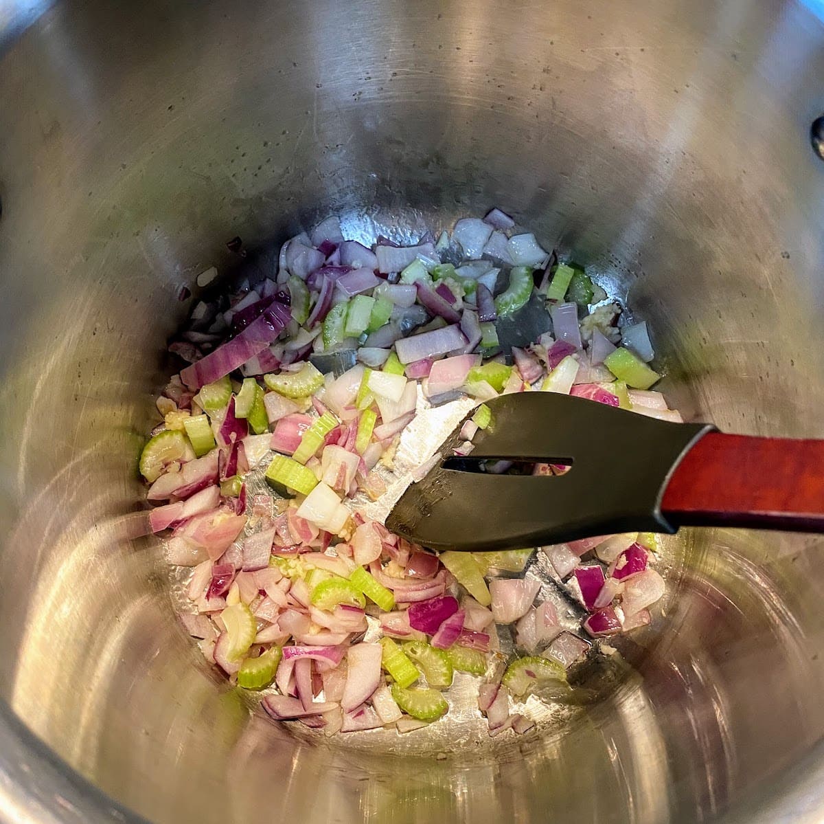 chopped onion, celery and garlic being stirred with a spatula in large, silver stockpot