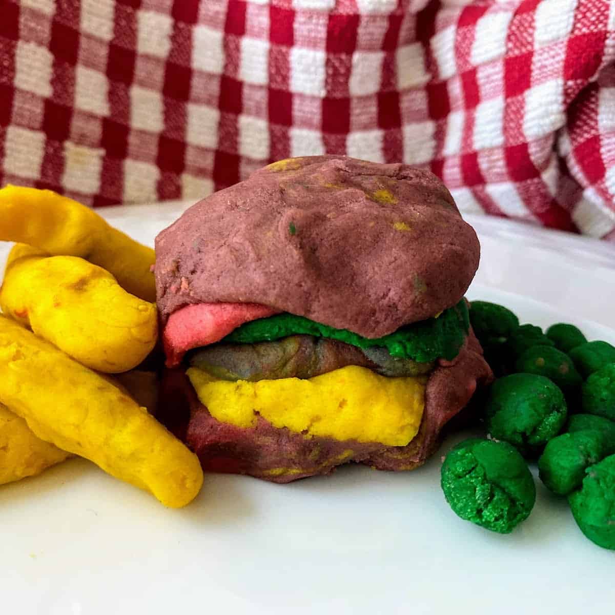 A burger, fries and peas made from playdough on a white plate. 