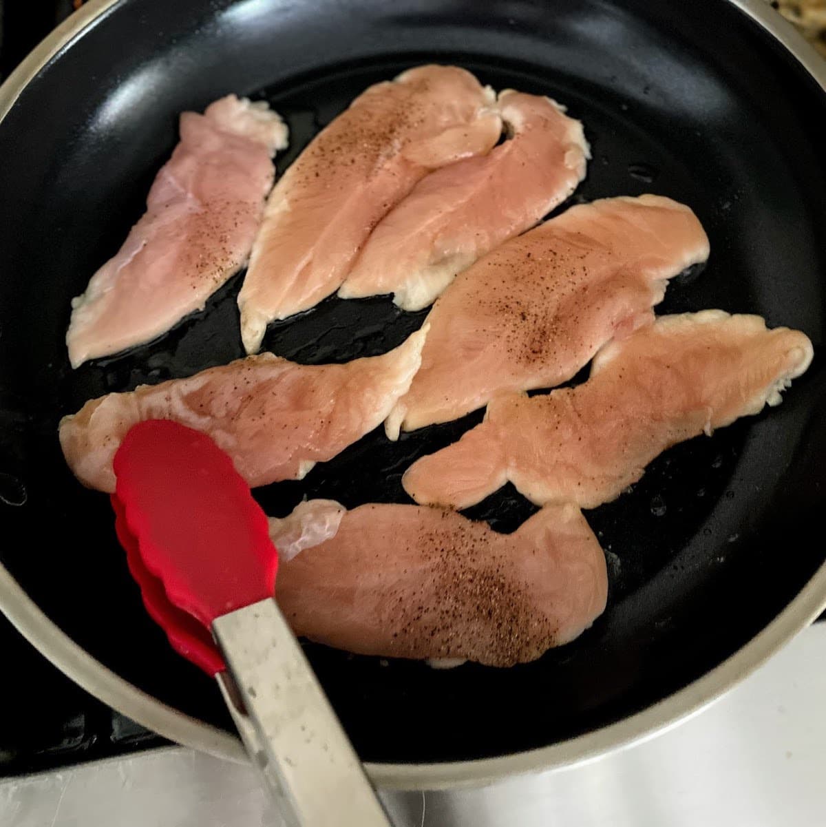 Hand holding tongs to turn over raw chicken strips in pan