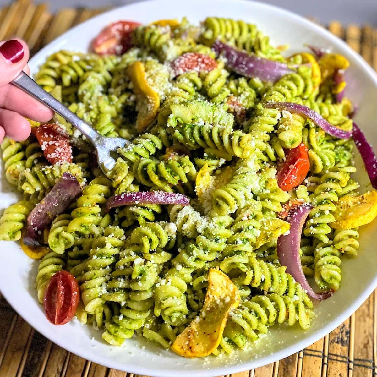 hand holding fork and bowl of pasta with roasted veggies and pesto sauce