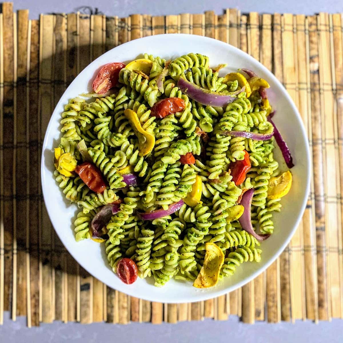 white bowl of pesto pasta, roasted tomatoes, squash and onion on bamboo placemat