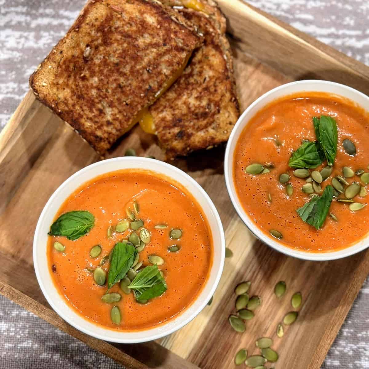 Two bowls of tomato soup topped with basil and served on a try with a side grilled cheese sandwich.