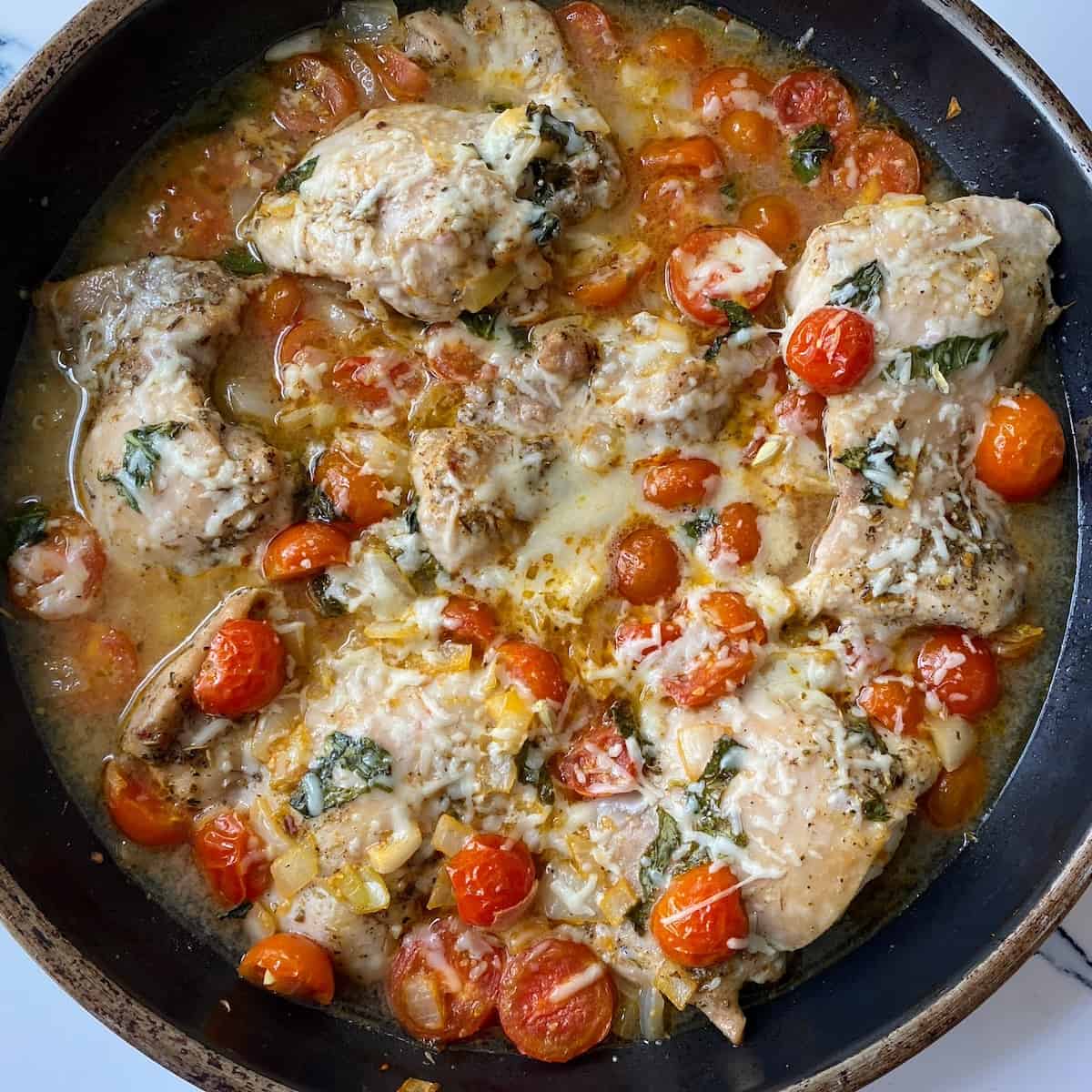 chicken pomodoro in black pan topped with basil, cheese and tomatoes.