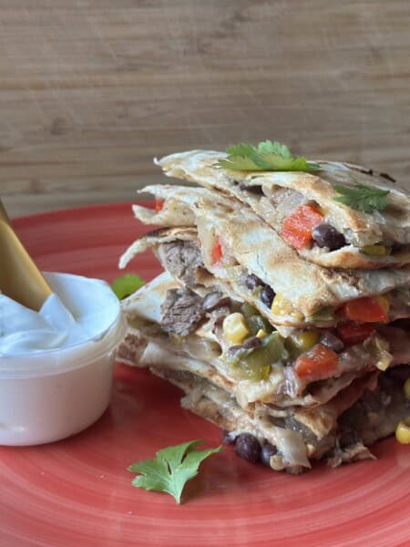 Stacked steak quesadillas served on a pink plate with a side of sour cream.