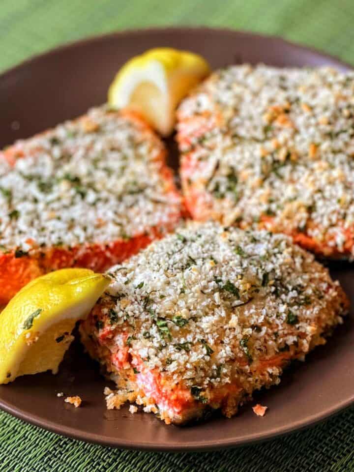 3 fillets of salmon encrusted with panko next to lemon wedges on brown plate on green placemat