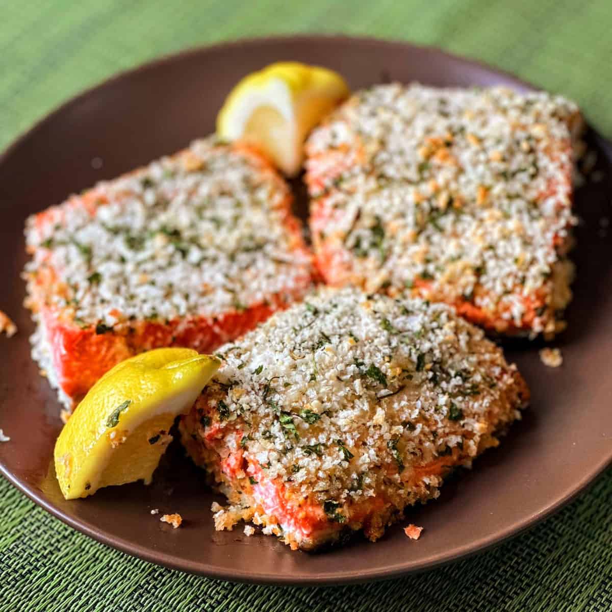 3 fillets of salmon encrusted with panko next to lemon wedges on brown plate on green placemat