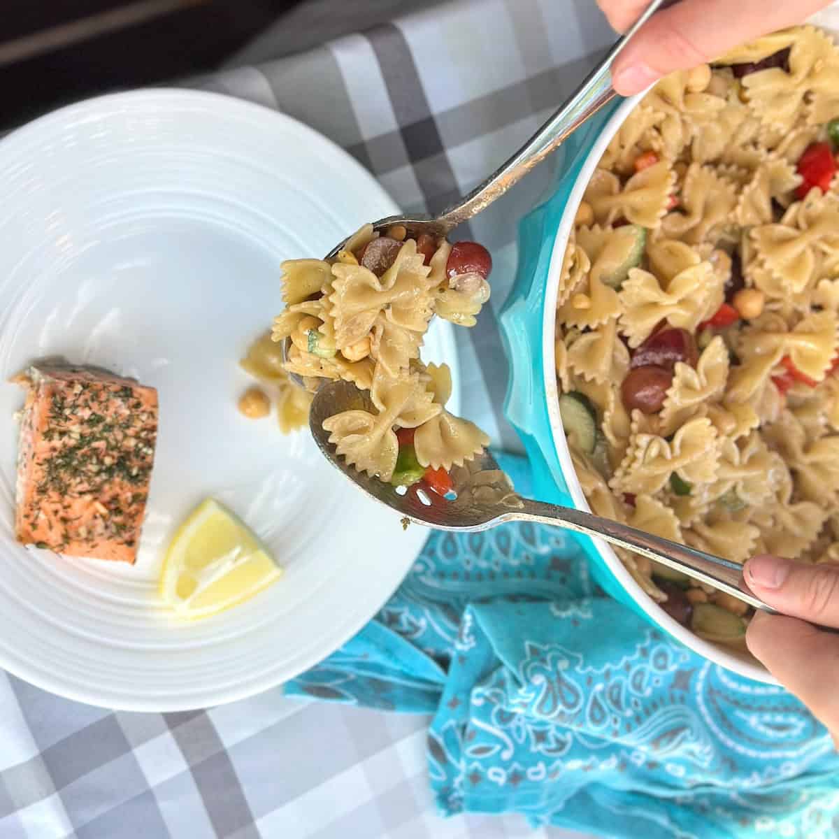 hands with silver spoons dishing pasta salad onto plate with salmon and lemon wedge