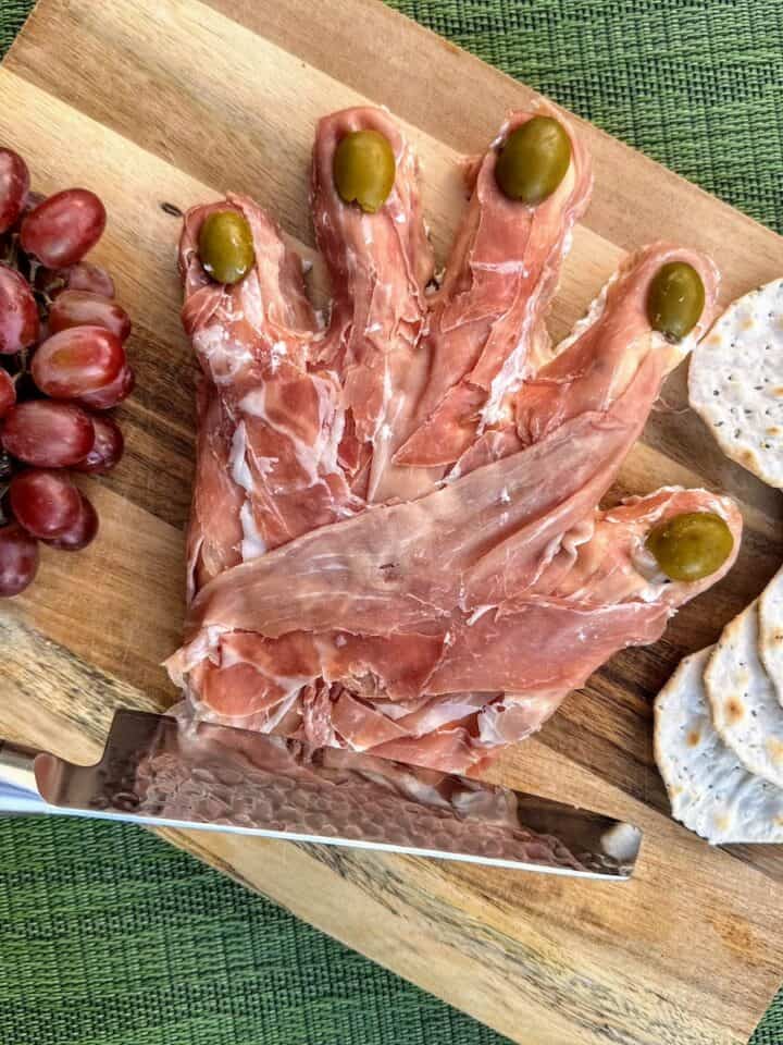 severed prosciutto hand on cutting board with grapes and sharp knife