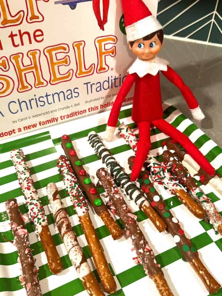 elf wearing hat and sitting on tray of christmas-decorated pretzel dessert rods