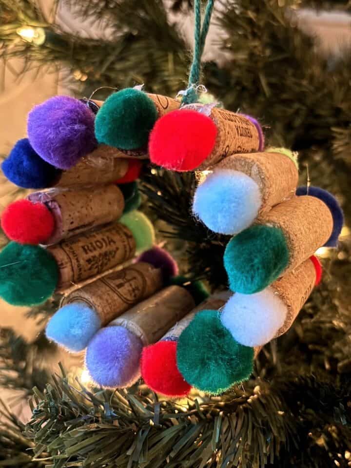 Wine cork ornament with colored pom-poms hanging from a Chistmas tree.