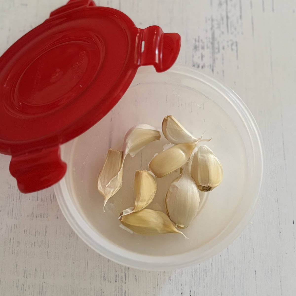 Several cloves of garlic in a Tupperware bowl with a red lid on the side. 