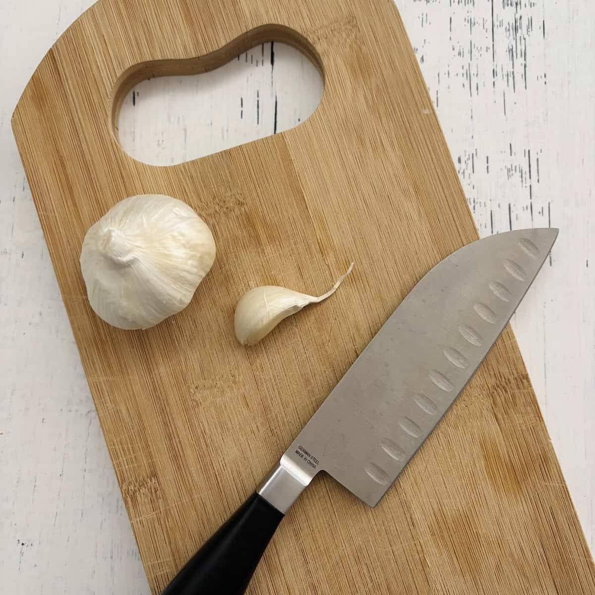 A chef's knife on cutting bar next to garlic clove to show the knife method for peeling garlic. 