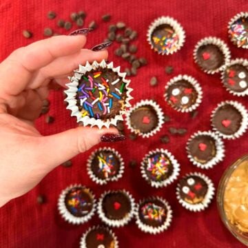 hand holding a peanut butter cup with rainbow sprinkles above a red tablecloth spread of more peanut butter cups, chocolate chips, and a cup of peanut butter