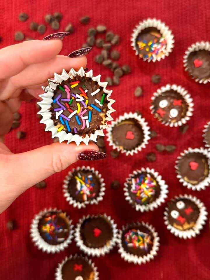 hand holding a peanut butter cup with rainbow sprinkles above a red tablecloth spread of more peanut butter cups, chocolate chips, and a cup of peanut butter