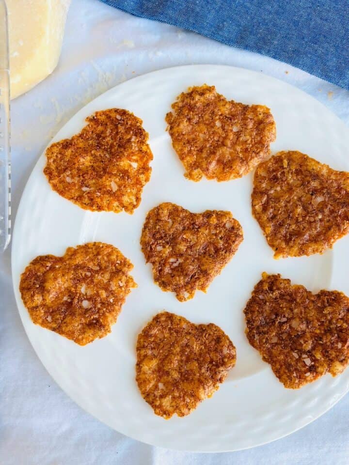 baked cheese crisps on a white plate surrounded by a cheese grater and block of parmesan cheese.