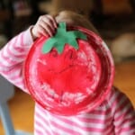 A young child holding a paper plate tomato over her face.