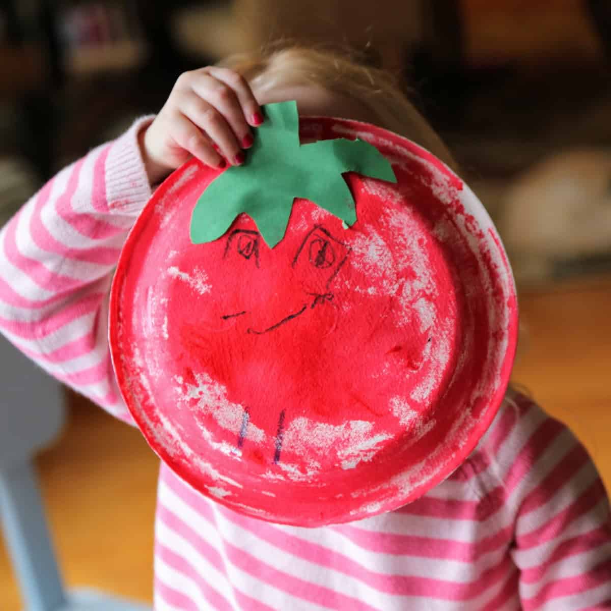 A young child holding a paper plate tomato over her face. 
