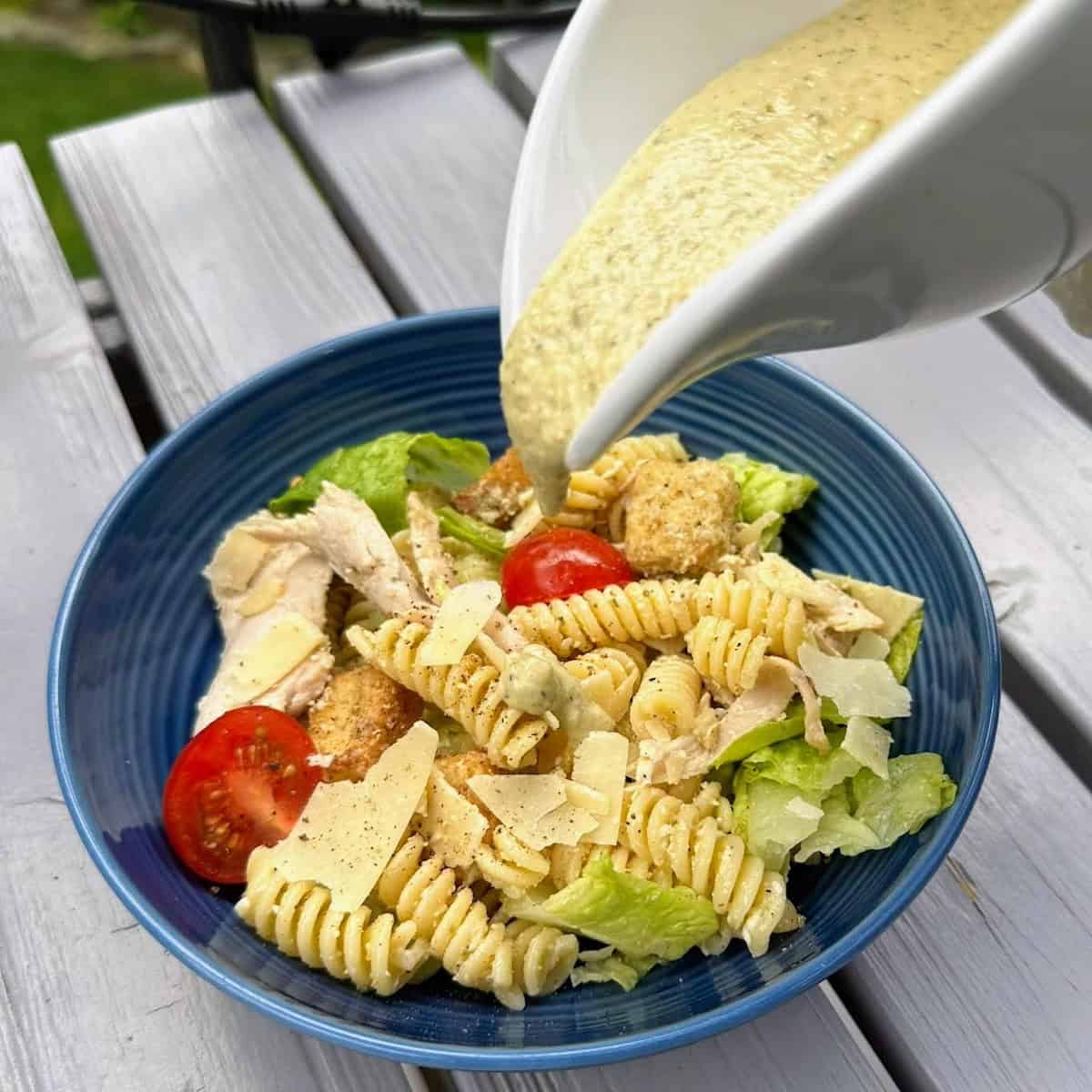 white sauceboat pouring dressing on top of salad in blue bowl on grey deck table