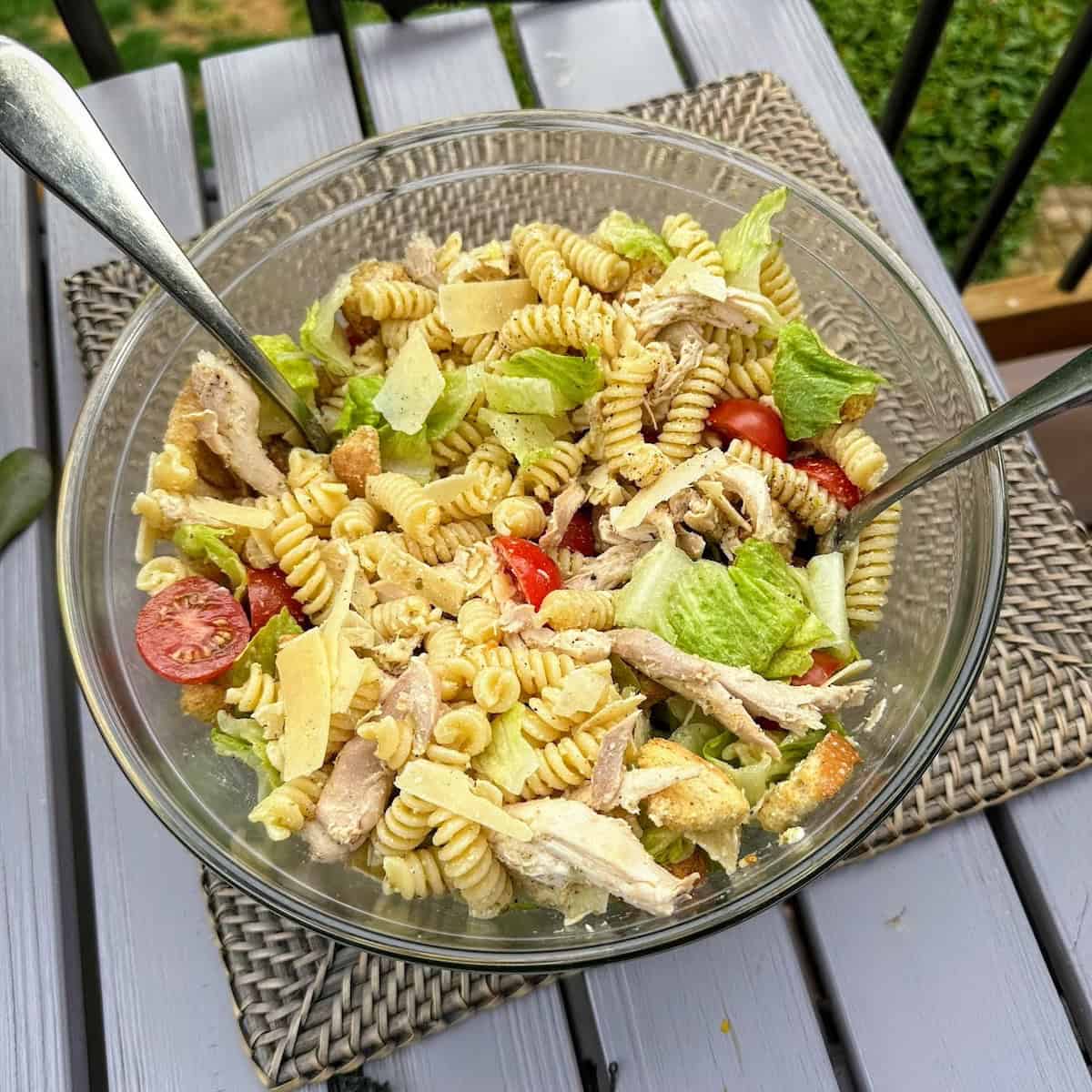glass Caesar pasta salad bowl with 2 silver serving spoons on brown woven placemat on grey deck table
