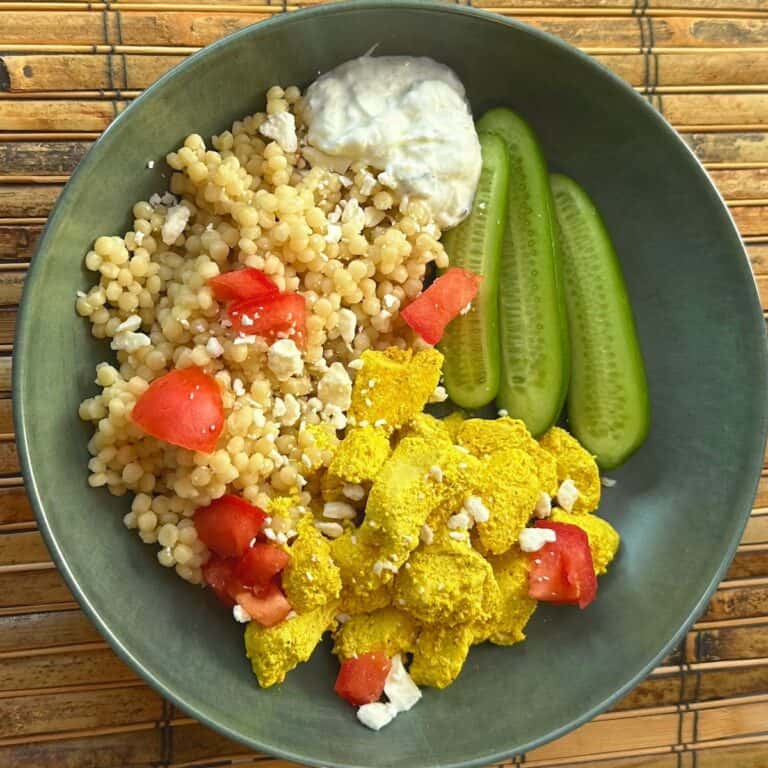 green plate with yellow Mediterranean chicken, cucumber, couscous, Tzatziki, feta and tomato