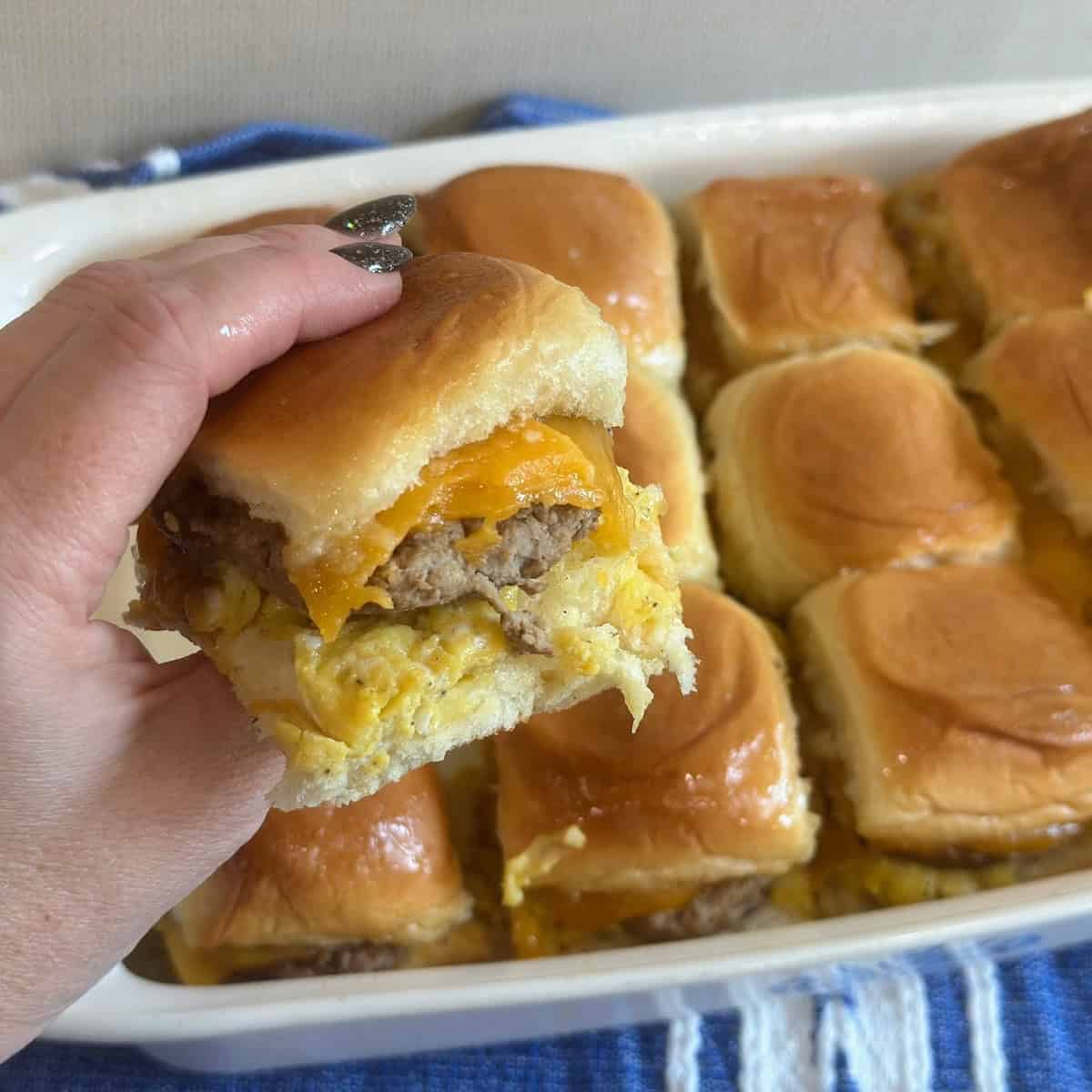 A hand holding a a breakfast slider over a casserole dish filled with sausage, egg and cheese sliders.