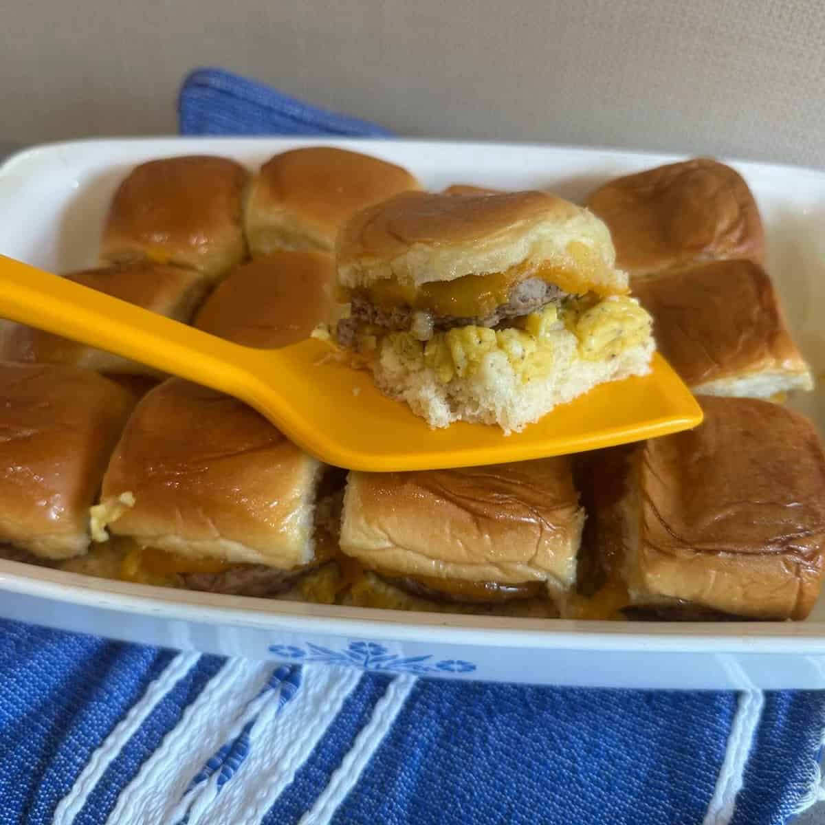Slider held in yellow spatula over casserole dish of sliders.