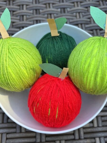 Four apple crafts made with colored yarn in a white bowl.