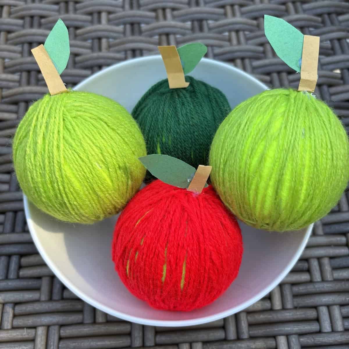 Four apple crafts made with colored yarn in a white bowl.