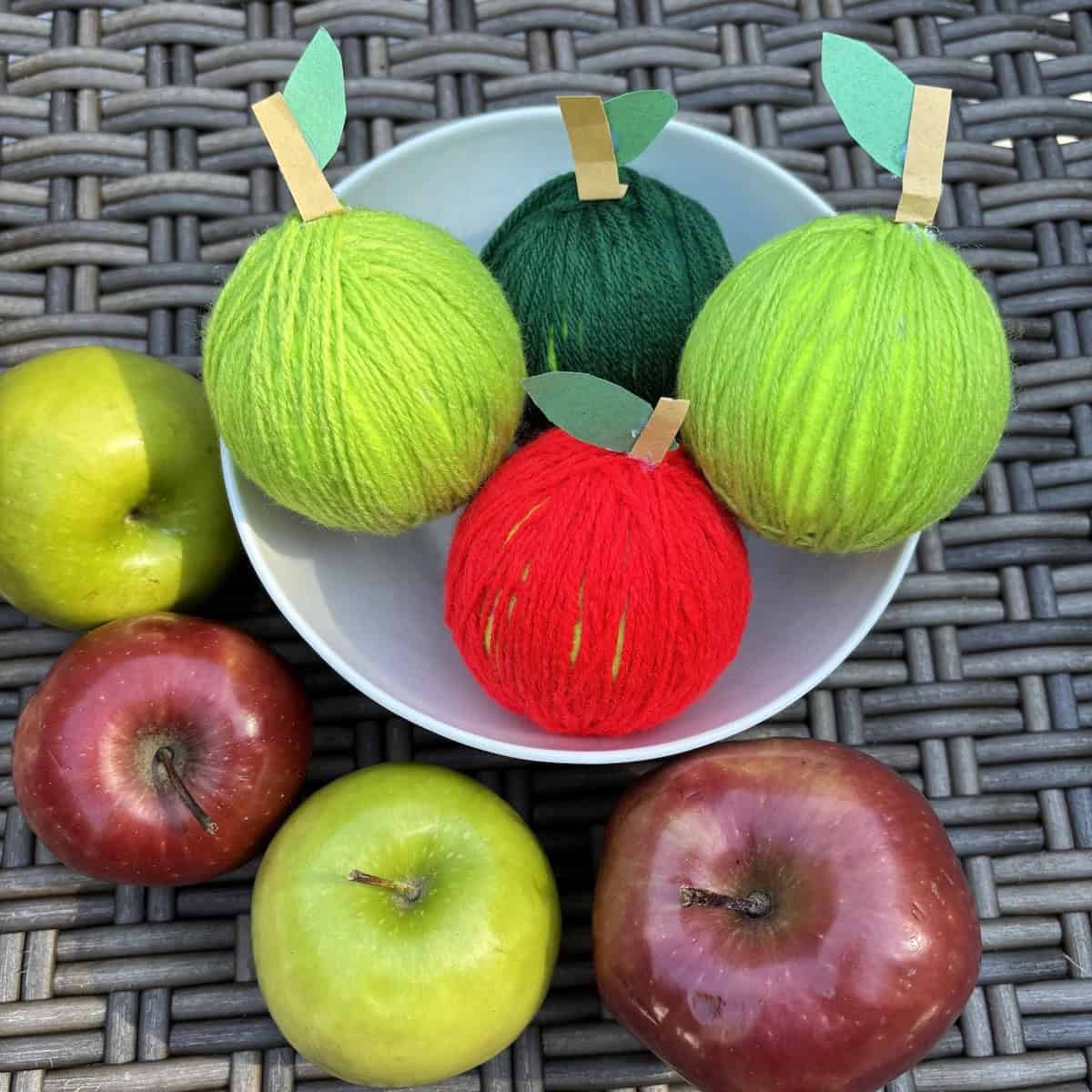 Four apple crafts made from tennis balls and yarn in a white bowl surrounded by real red and green apples.