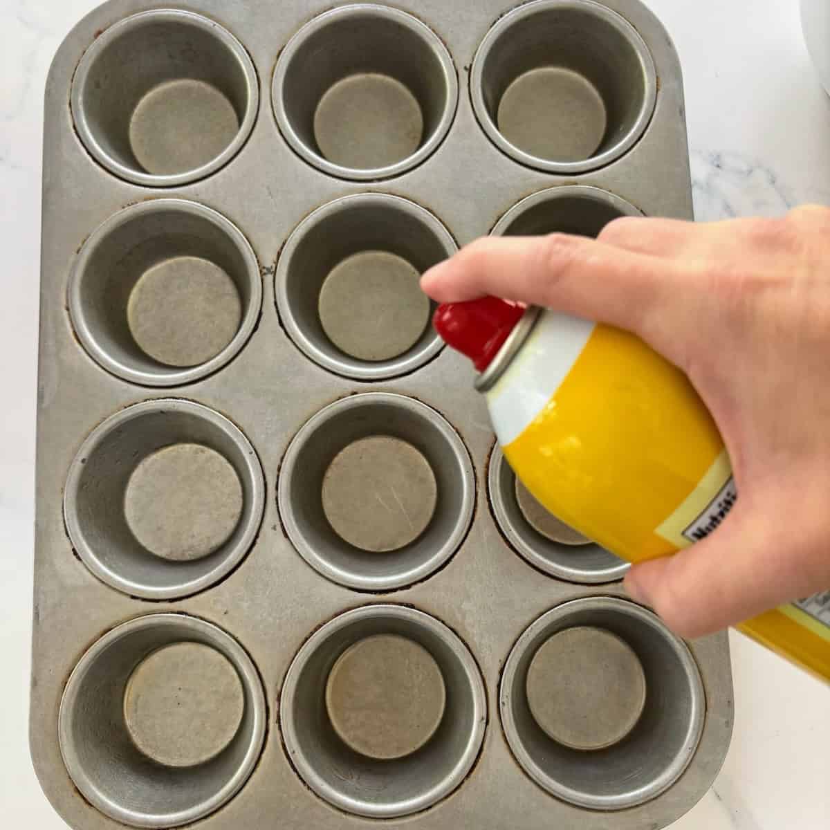 Person spraying non-stick spray into muffin tin for baking preparation.
